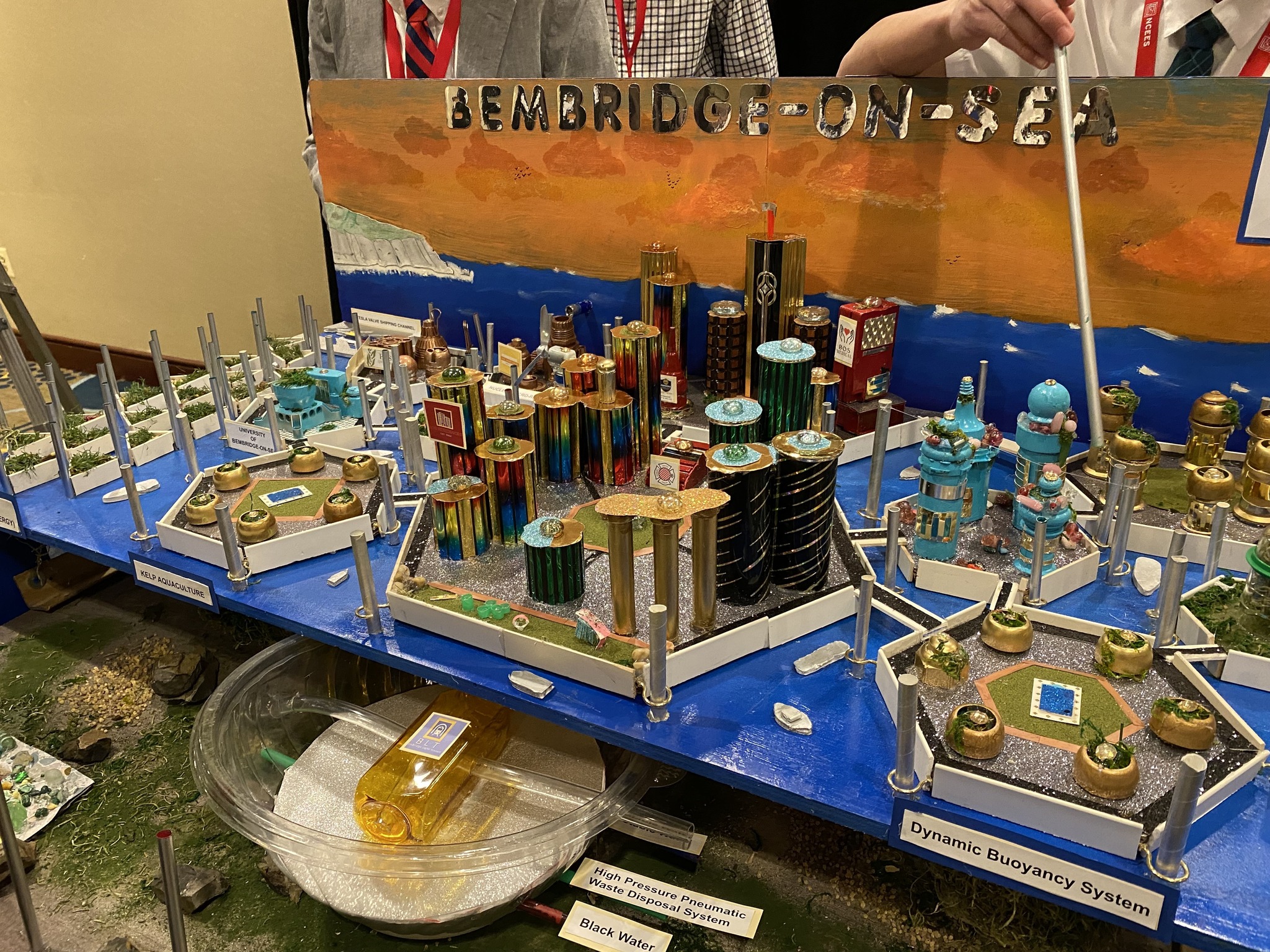 Future City Competition entry titled 'Bembridge-on-Sea,' showcasing a detailed model of a futuristic coastal city. The design features innovative engineering concepts, including a dynamic buoyancy system, high-pressure waste disposal, and kelp aquaculture. The model is constructed with colorful materials, representing sustainable urban planning. Participants in suits and red lanyards present the project with a pointer, emphasizing key features.