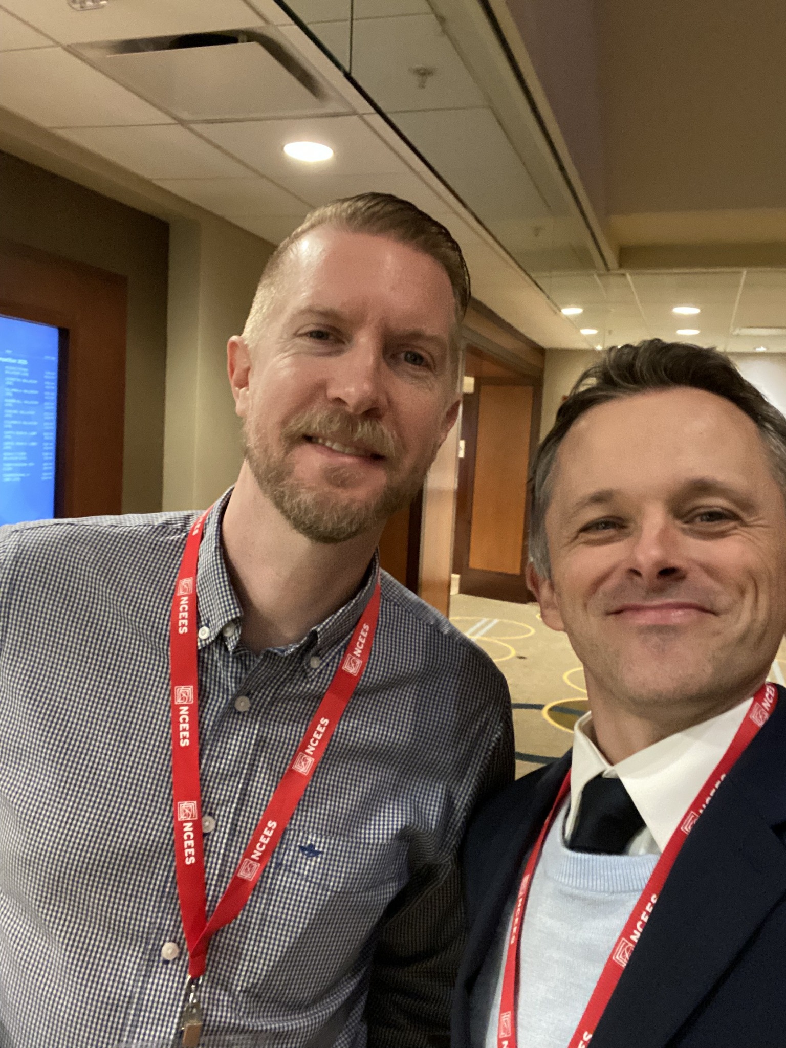 Two men at an NCEES conference taking a selfie, both wearing red NCEES lanyards. One is in a checkered shirt, the other in a suit with a light blue sweater. They are smiling in a professional indoor setting with a hallway and event signage in the background.