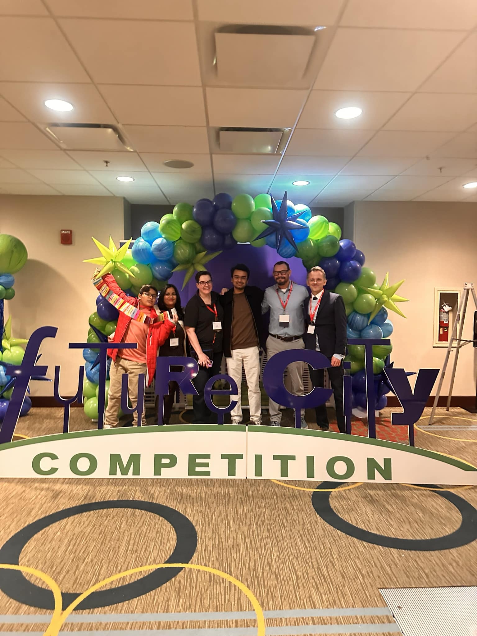 A group of Future City Competition judges stand together in front of a colorful balloon arch at the event. The large 'Future City Competition' sign is prominently displayed, highlighting the STEM-based competition focused on engineering and city planning. Attendees are wearing event badges and lanyards, showcasing a professional yet celebratory atmosphere.