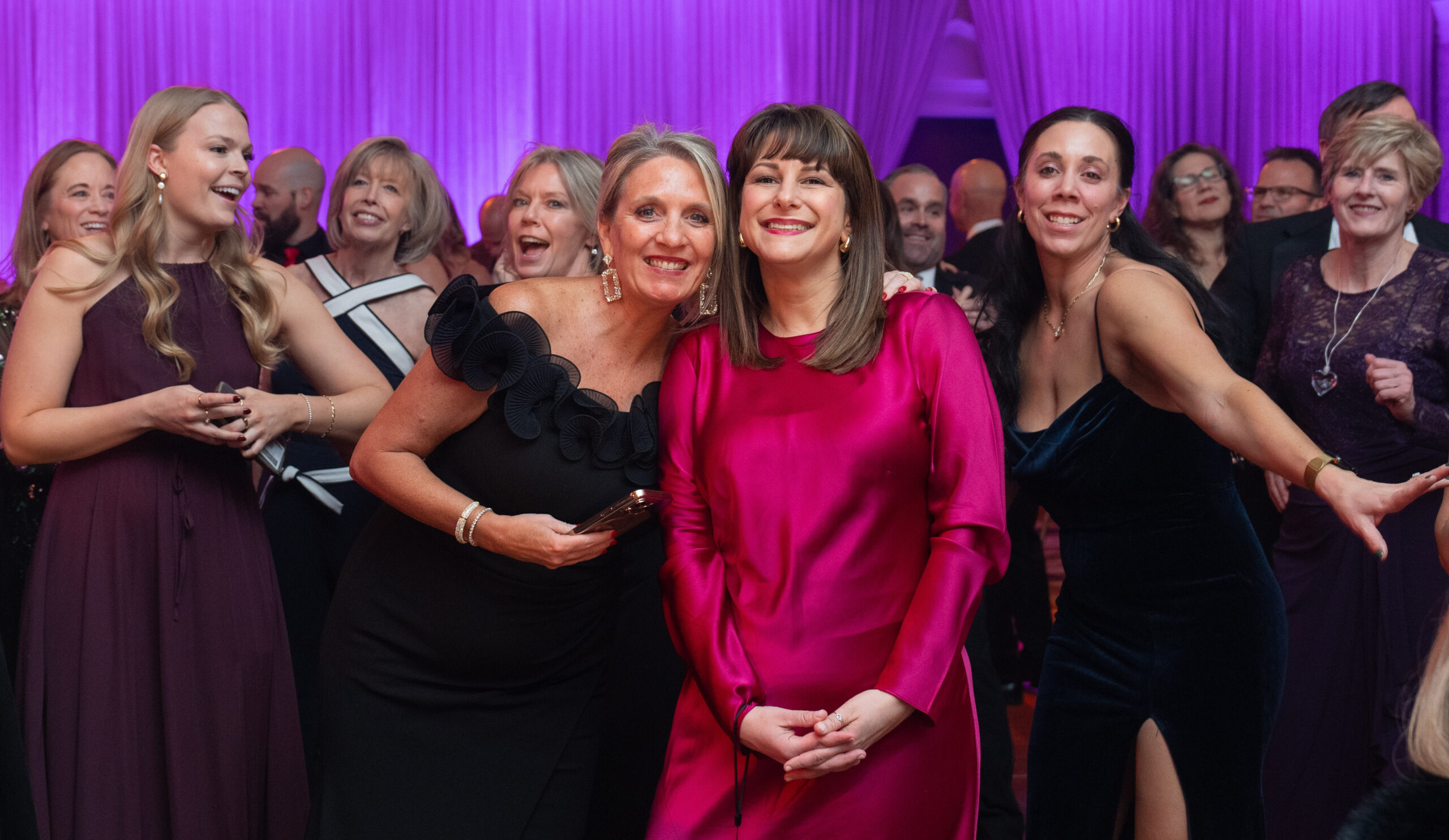 A group of elegantly dressed people enjoy a lively moment at a gala event, with purple drapery in the background. Two women in the center smile